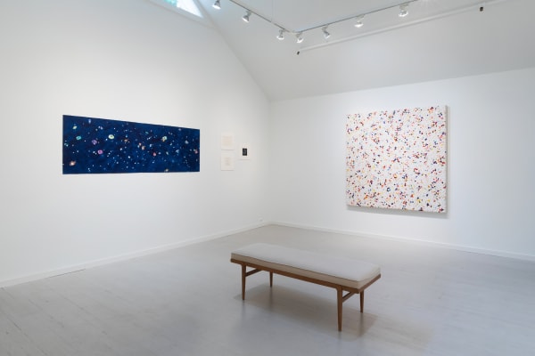 Two larger paintings by John Torreano, one a combination of dots in the shape of a square, and one a long rectangular starry procession of gems, hung on adjacent walls, with a bench placed in front.