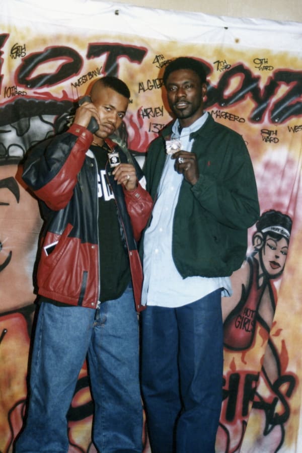 A photo by Polo Silk of himself and Juvenile standing in front of a Hot Boys airbrushed backdrop. Both hold their diamond pendants up to for camera. Juvenile is holding a 1990s cell phone to his ear.