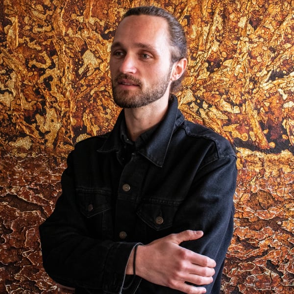 A thin white man cropped at the waist, hair pulled back, wearing a black Demin jacket, stands in front of a painted field of metallic gold and bronze