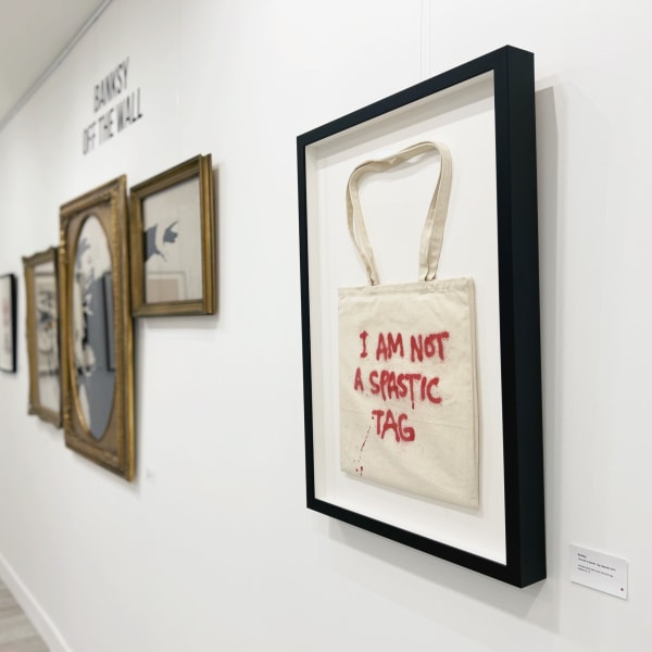 Banksy's 'I Am Not A Spastic Tag' artwork displayed in a gallery with other framed pieces on the wall.