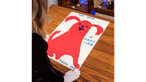 Person viewing David Shrigley's artwork 'No One is Happier Than Me' featuring a red cartoon-like creature on a wooden table.
