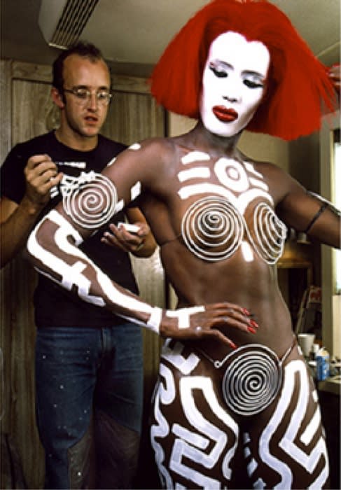 Color photograph of Keith Haring painting white graphic lines on the body of Grace Jones who is wearing a bright wig