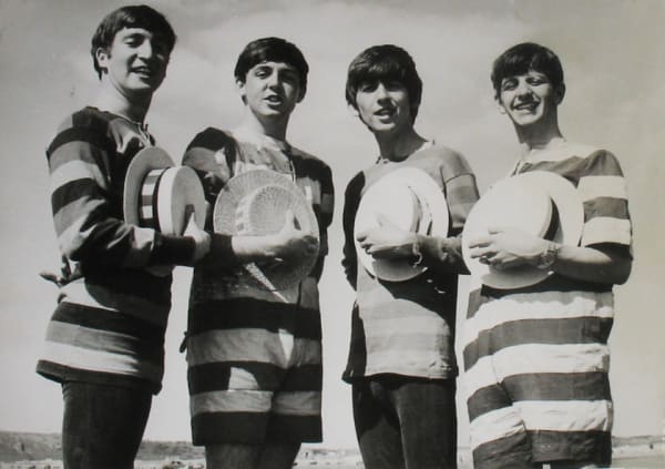 Black and white photograph of the Beatles on the beach in stripped shirts holding straw hats