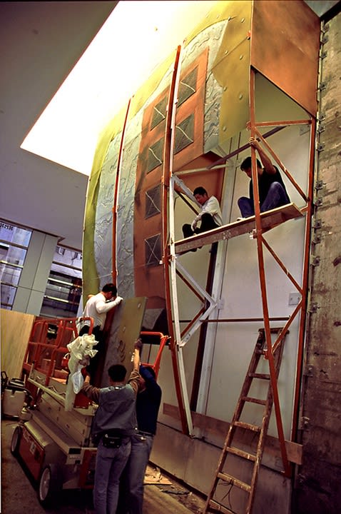 A crew of workmen build a large sculpture of dichroic glass by Warren Carther that appears as a curved opaque sail rising from the surrounding wall