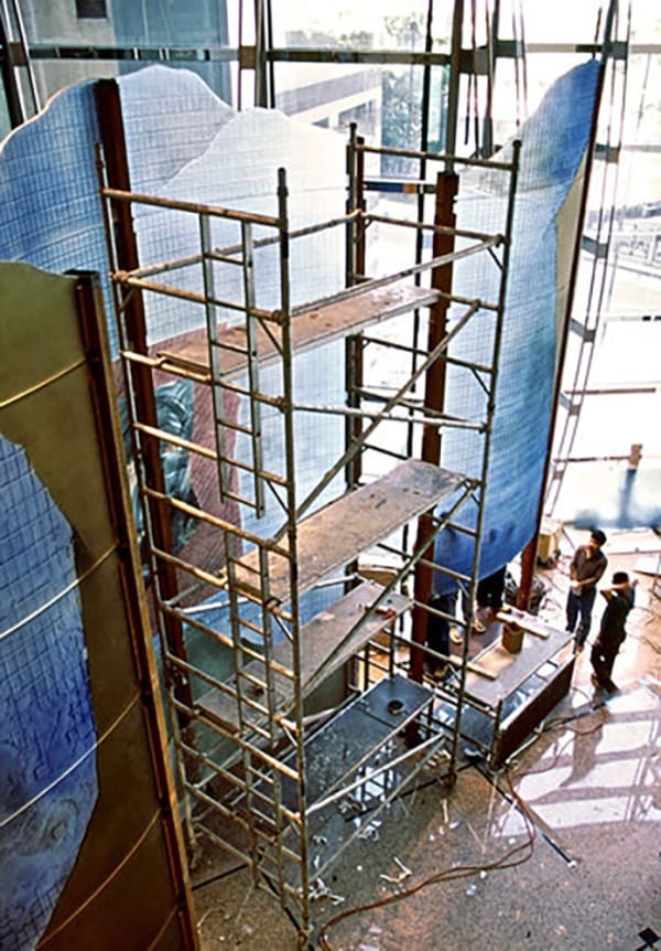 Large dichroic glass sculpture like an oversize modular screen (green, yellow, blue) with a construction scaffolding and men working