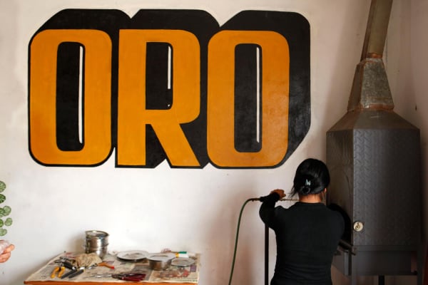 A woman stands away from camera in a gold and mining supply shop; the word for gold, 'ORO,' is painted on the wall
