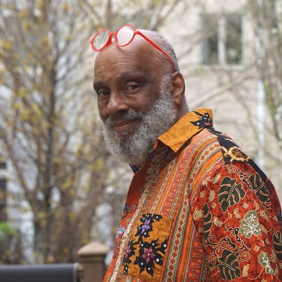 Portrait of artist Danny Simmons in an African textile shirt and bright orange glasses standing outside smiling at the camera