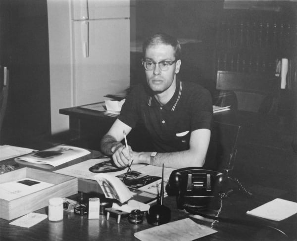 A man sits at a desk holding a pencil with a landline phone and papers neatly laid out before him