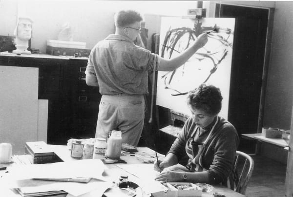 In an art studio at the Brooklyn Museum, a seated woman works paints at a desk and James Juthstrom stands painting at easel