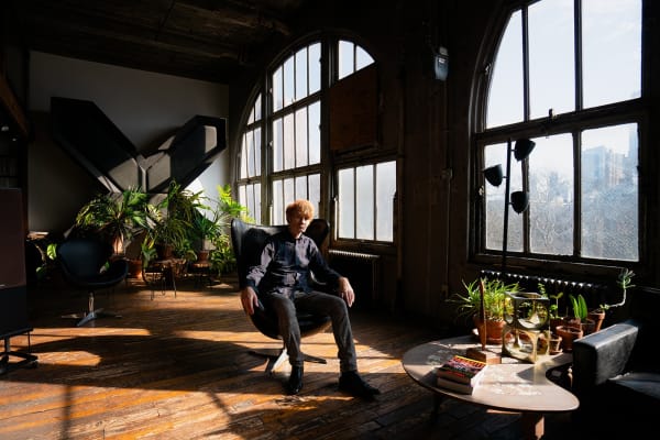 An artist, JG Thirlwell, sitting in a chair amidst potted plants next to large arched windows in an industrial loft setting