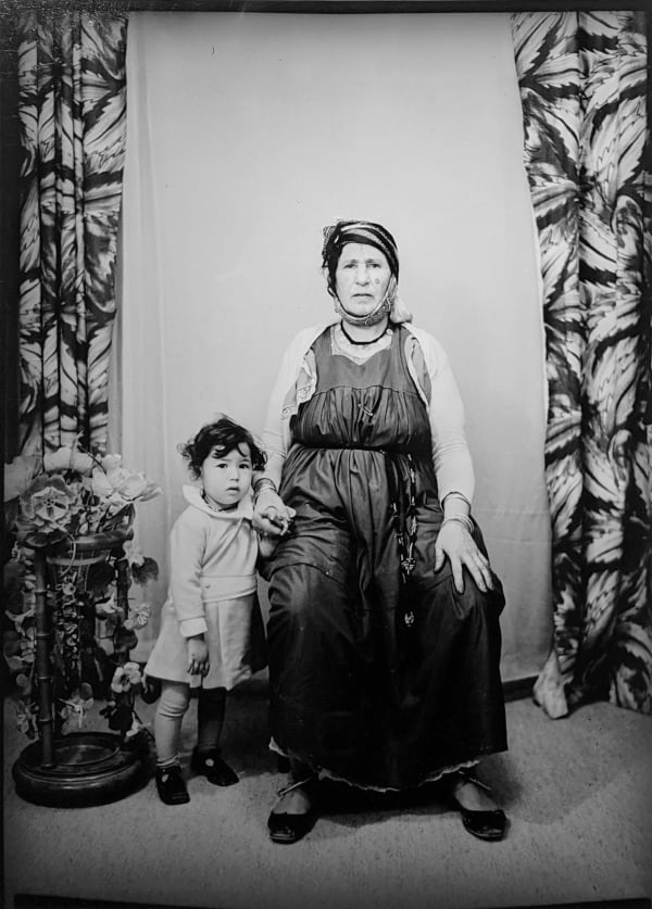 1960s black and white photograph of a older woman sitting down in a dress with a headwrap with a young boy to her left holding her hand