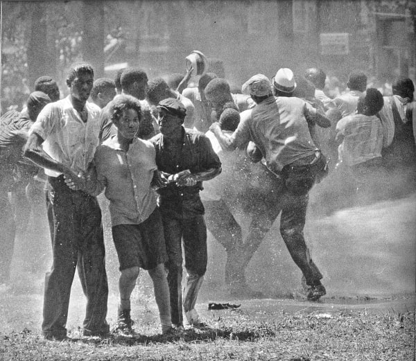 Black and white photograph of protesters in Kelly Ingram Park in Birmingham, Alabama being sprayed with fire hoses