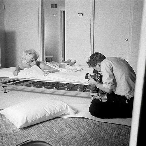 Marilyn Monroe lays on a bed amidst white sheets while Douglas Kirkland kneels and takes her photograph
