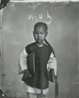 Period photograph of Chinese schoolboy with a long plaited braid wearing the traditional outfit and a book under his arm