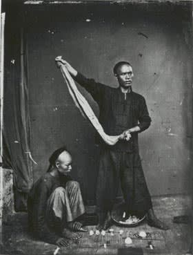 Period photograph of two Asian men in streetwear (one crouching, the other, with long queue, rolling dice in a scarf)