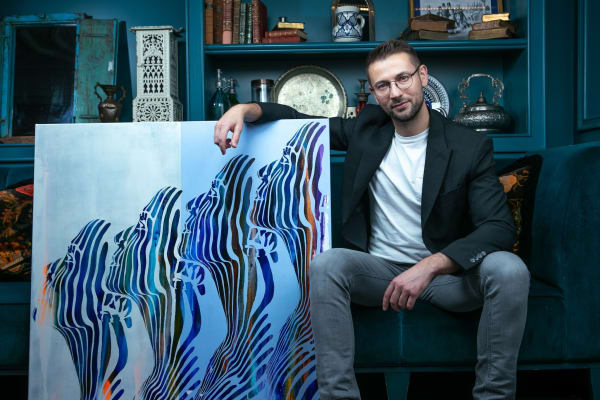 A person posing with a vibrant, abstract painting featuring a series of stylised faces in blue and white tones, set against a richly decorated interior.