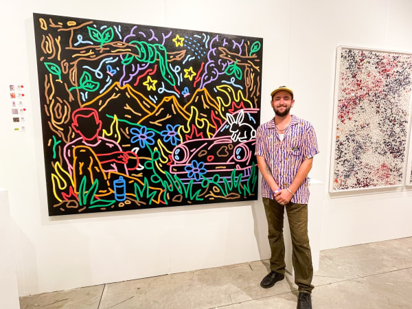 Man posing in front of paintings displayed at Carousel Fine Art's booth at the art fair, Context Art Miami 2021.