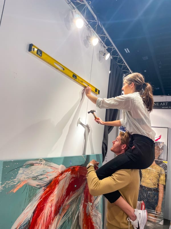 Woman on man's shoulders hanging a painting at Carousel Fine Art's booth at the Palm Beach Show art fair, 2022.