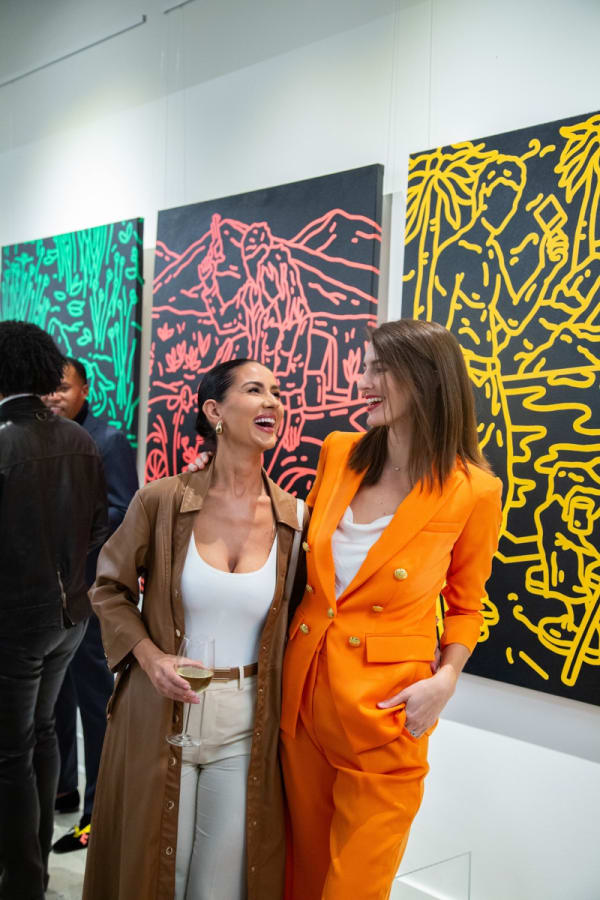 Women laughing in front of paintings displayed at Carousel Fine Art's new gallery space in Buckhead Village, Atlanta during an event for their grand opening.