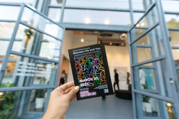Hand holding exhibition booklet in front of Carousel Fine Art's gallery space in Miami, FL for Chance Cooper's Spotlight Exhibition, "Between Me and You."
