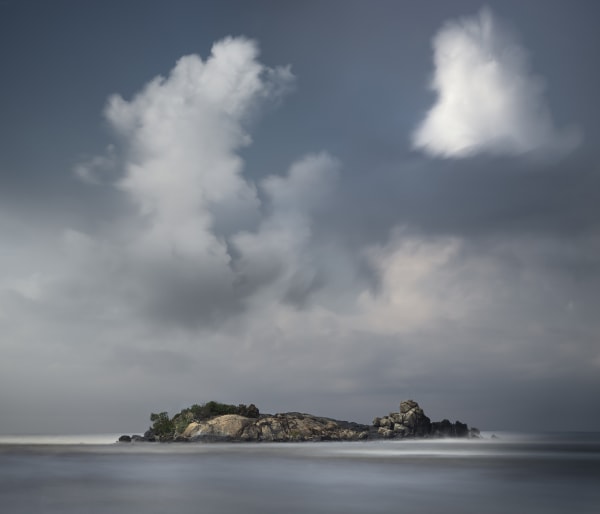 small island in middle of tranquil waters with puffs of clouds on horizon.