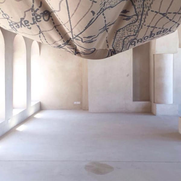 PORTRAIT OF A LAKE 2017, print on natural fabric (300x400cm), hemp strings, 3L of water produced by a nearby dehumidifier, poured every day Installation view from Sharjah Biennial 13 Tamawuj, curated by Christine Tohme, Sharjah, UAE Portrait of a Lake consists in a suspended piece of map. It shows us the outlines of a lake and its surrounding grounds, now hanging above us. As water pours on it, the representation of the lake, dating back from 1938, becomes humid again; the lake is reactivated. As it crosses the representation of the lake, water becomes water from the lake. Dripping down onto the floor, it slowly draws the outlines of the lake anew; A self-portrait, drawn by the lake itself. The portrayed lake is Yammoune – Little Sea (from Ugaritic, "yam"). On its West shore are scattered the ruins of a Roman sanctuary. Dedicated to Astarte-Venus, goddess of fertility, the sanctuary also held statues of shepherd-gods protecting pastoral life and agriculture. Built next to Nabaa el-Arbain, the main spring flowing into the lake, the sanctuary had a direct connection with water and purification rituals. Abundant with water from the spring at snowmelt, Yammoune Lake progressively empties, until the end of summer, forming a fertile depression. Culminating above the fertile valley of the Bekaa, which it partly irrigates, its water also irrigates the illegal crops cultivated in the village of Yammoune. Situated on the Levant geological fault, marking the boundary between the eastern Mediterranean and the Arabian plates, Yammoune Lake is a “pull-apart”: a void constituted by the slippage of the two tectonic plates. The fault, departing from Jordan, crosses Lebanon’s entire geography, as well as Israel and Syria, before ending in Turkey; Divided insegments, breaking one by one, at different times, it is responsible for a significant amount of seismic events in the Middle-East. Closely following the nature, cycle and history of the lake, the installation is also reminiscent of a sail, a tent or a breast.