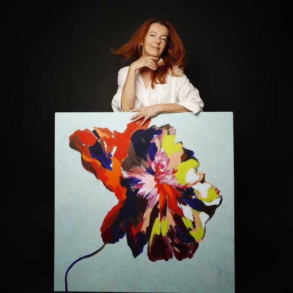 Portrait of artist Caroline Muscat standing behind one of her paintings, featuring a bold, expressive flower form in vivid reds, blues, yellows and pinks against a pale blue background.