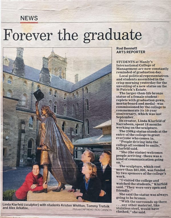A newspaper clipping titled "Forever the graduate" by Rod Bennett from the Manly Daily. The article features a photograph of sculptor Linda Klarfeld with students Kristen Whitton, Tommy Trettvik, and Alex Arkatov at the unveiling of a larger-than-life bro