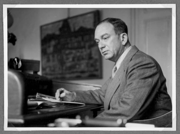 A black and white photograph of Otto Kallir: middle-aged caucasian man in a suit seated at a desk looking off to the left. An oil paintings hangs on the wall in the background