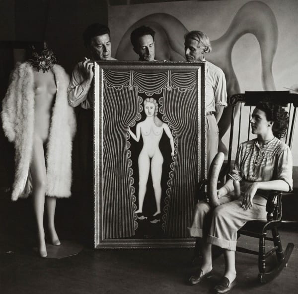 A black and white photograph of three clothed men standing behind an oil painting of a nude woman done in a folk-art style. A semi-nude model stands at the left of the painting while a clothed woman sits in a chair to the right