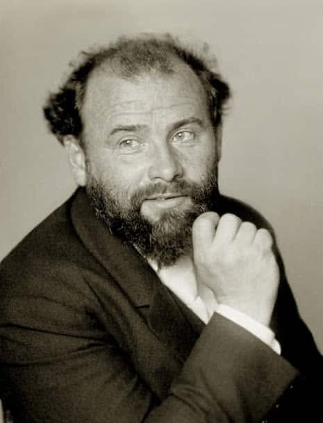 A black and white photograph of Gustav Klimt: a middle-aged, balding white male in a suit, his right hand lifted to his chin as he looks off into the distance