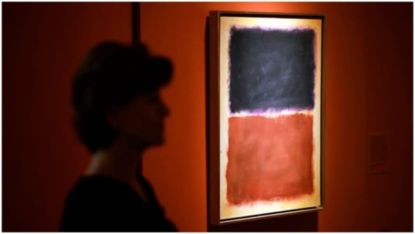 The silhouette of a person stands at left before an illuminated abstract painting in shades of black, red and beige hanging on a red wall