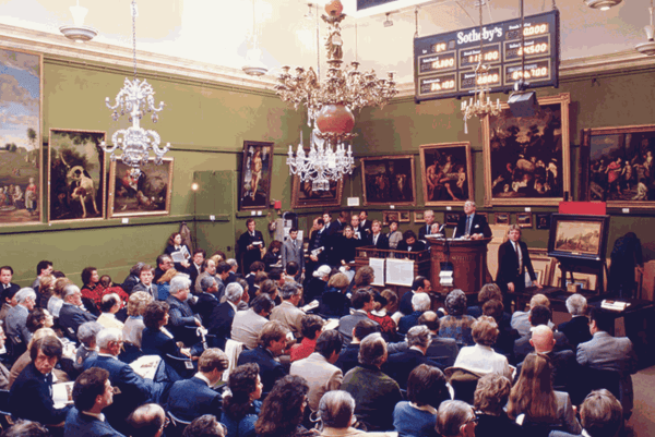A color photograph showing an elevated view of an auction room. An auctioneer stands at a podium before a room of seated individuals, oil paintings adorning a green-walled room with multiple chandeliers hanging down
