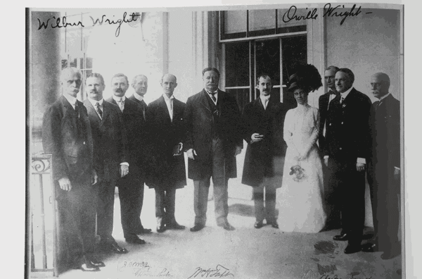 A black and white photograph from a book showing 10 men and 1 woman standing together. Among the group are Wilbur Wright, Orville Wright and Theodore Roosevelt.