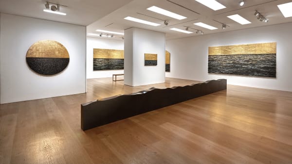 Installation view of Yoan Capote's exhibition in a white walled gallery featuring black and gold paintings and a long low black stone sculpture