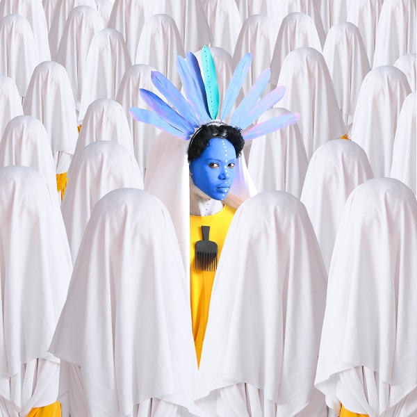 A woman with blue-painted face, black hair, and a blue and green feathered headdress stands in the middle of a group of figures shrouded, ghost-like, in white sheets.