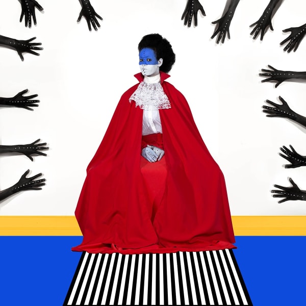 Seated portrait of a black-haired woman in a red cape and white lace collar, her face painted blue on top and white on the bottom, against a white background, with dark black hands reaching into the frame from the top and both sides.