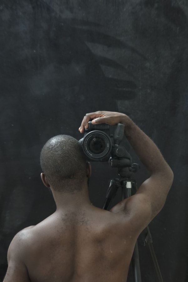 Half-length portrait of a shirtless Black man, seen from behind, with his right arm raised holding a camera against his head, its lens aimed at the viewer.