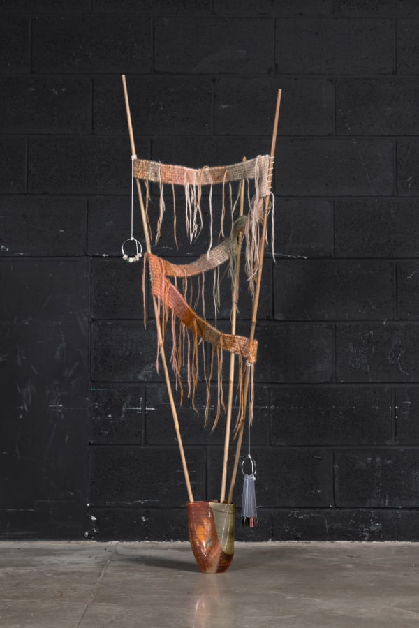 A hand made ceramic vessel with three bamboo sticks coming out of the three openings at the top. The sticks have a hand knit textile zigzagging between them. From the top of one bamboo stick hangs a key ring with plastic beads on it. From near the bottom