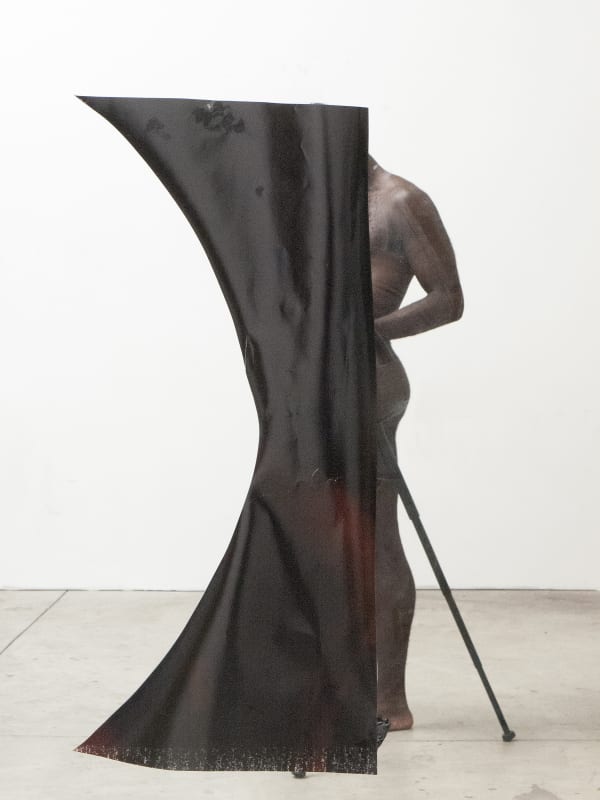A nude photographer stands in profile in a studio, his back half visible, as is one leg of his tripod. His front half is concealed behind a large piece of shiny black material.