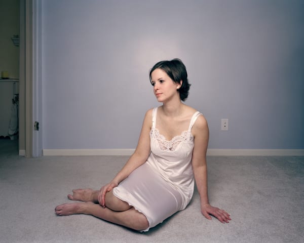 A woman in a white slip seated on the floor of a bare, white-carpeted room, her bare legs revealing amputated toes on both her feet.