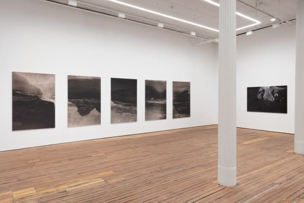 A series of large black and white photographs depicting images of water are hung in a line in the middle of a wall to the left. This photo looks to a corner of the gallery where that wall is visible, along with a column, wood floor, and another gallery wa