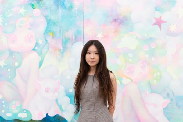 So Youn Lee stands in front of two of her large pastel colored paintings in a white wall art gallery.