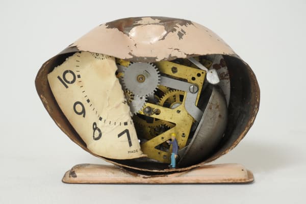 A smashed metal alarm clock with a small figurine of a janitor sweeping at the bottom.