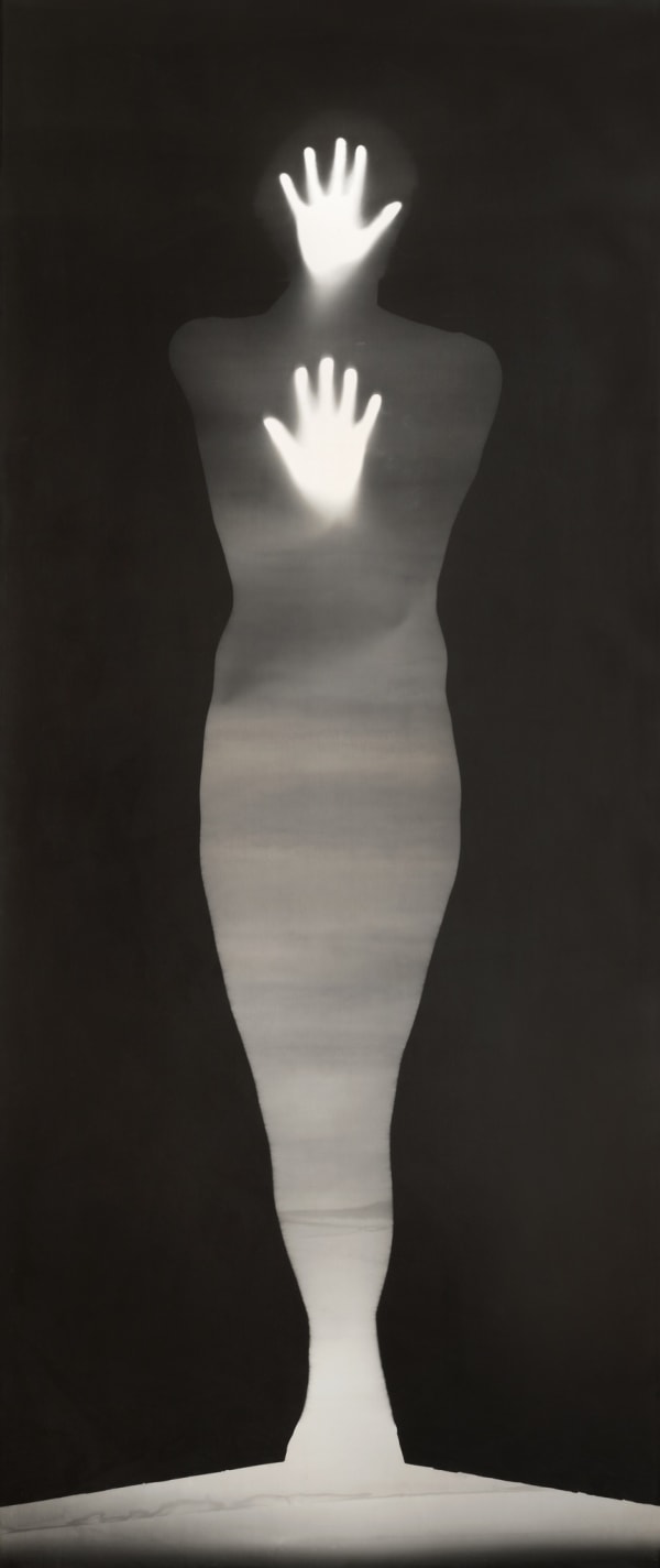 This image features an artistic black-and-white photograph or artwork with high contrast, creating a silhouette effect. A human figure appears to be at the center, surrounded by darkness. The body of the figure is mostly obscured except for two glowing, bright areas that resemble hands with fingers spread out, positioned one above the head and the other at chest level. The silhouette seems to be standing upright, with the light creating an ethereal or supernatural impression. The background is uniformly dark, enhancing the luminosity and focus on the highlighted hands.