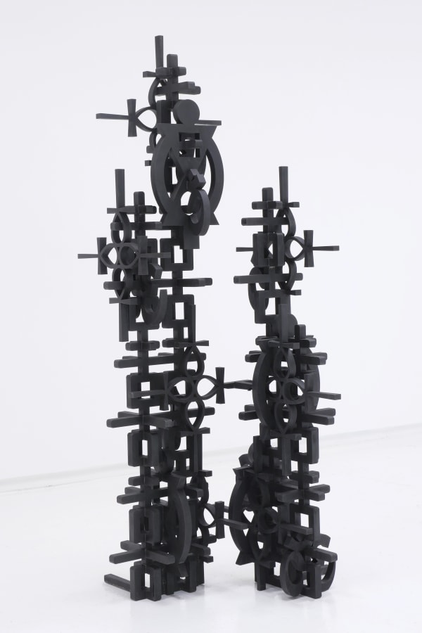 This image shows a sculpture composed of numerous black geometric shapes interconnected to form two vertical, symmetrical structures. The shapes resemble mechanical parts, such as gears, cogs, and crossbeams, intricately assembled into a complex form. The