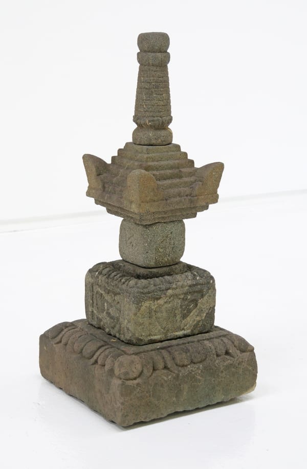 The image shows a traditional stone pagoda sculpture often found in East Asian gardens. The pagoda is constructed with multiple layers, each with its own distinct shape and texture, contributing to the overall tiered design. From the bottom, there's a bro