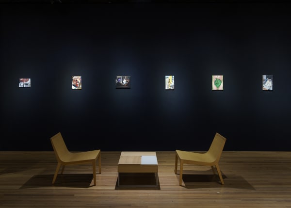 Small oil paintings in an art fair booth against a black wall illuminated by spotlight and two chairs.