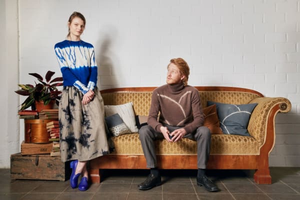 a man sitting on a tan couch looking at a woman sitting on the arm of the couch. There's a stack of trunks next to the couch.