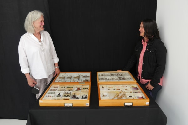 Isabella Kirkland and Lauren Esposito flank table covered with four wooden boxes displaying dead insect specimens.